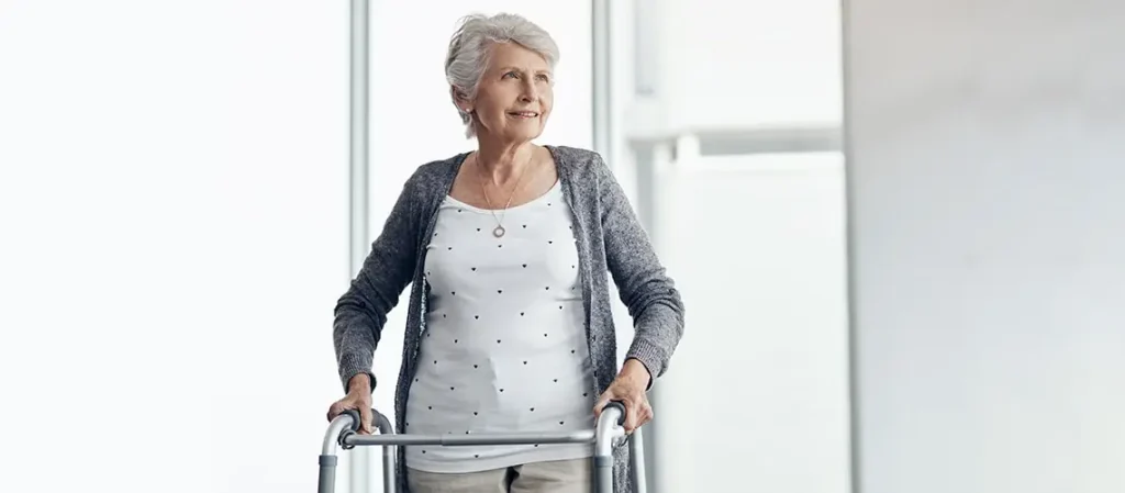 A smiling senior woman in a gray sweater walking with a short walker. For seniors, walkers are often critical.