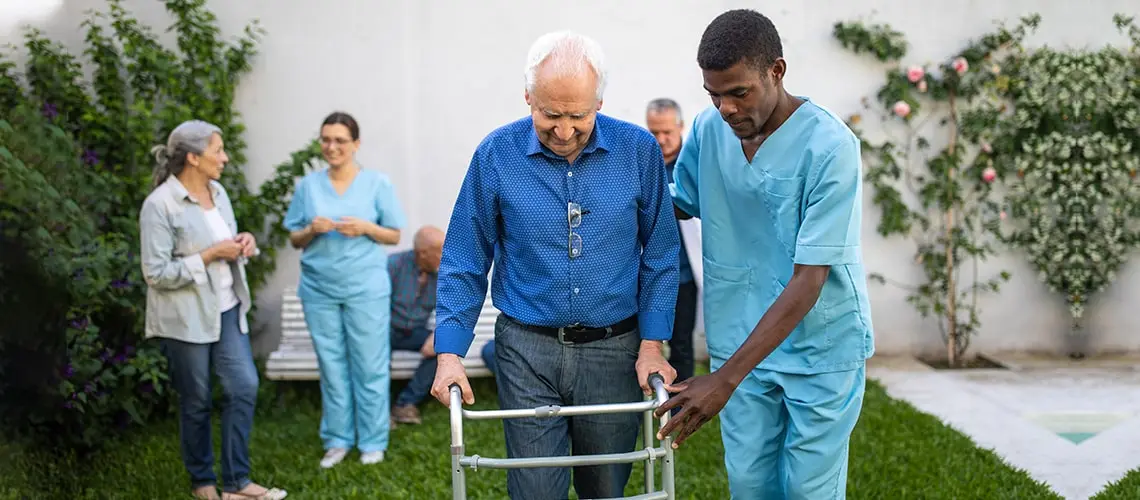 A male nurse supporting a senior man while he is walking with assistance of narrow walkers for seniors.
