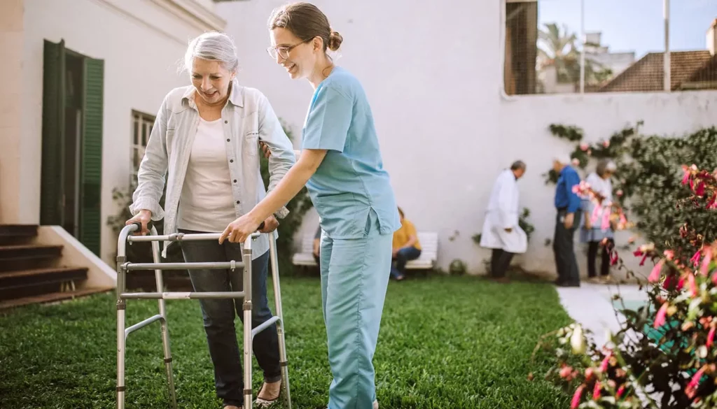 Standard walkers are also known as walkers without wheels. A senior woman walking with the help of a standard walker with a female nurse by her side.