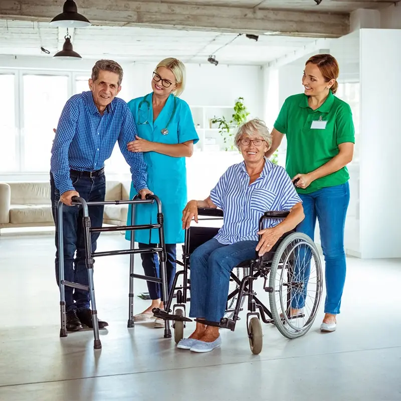 A senior man using a standard walker and a senior woman in a wheelchair. A female doctor and a female nurse are assisting them.