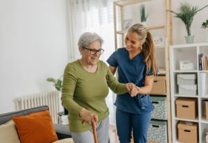 Female caregiver helping and supporting senior patient to walk at home, senior woman is standing and holding walking cane.