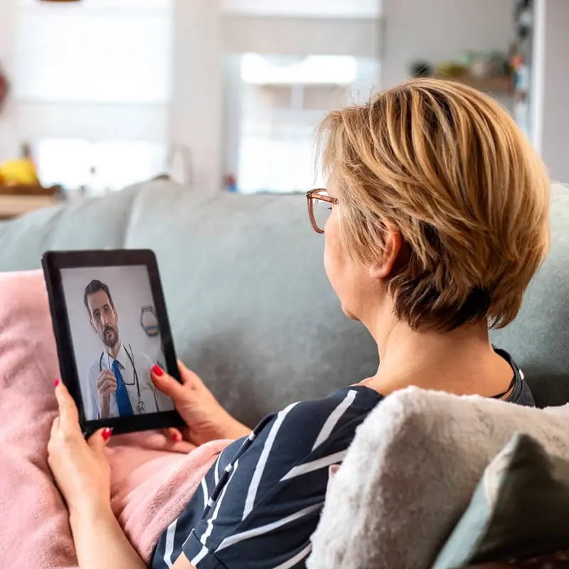 One of the advantages of accessible home health care is the freedom to get consistent care at your own home. A senior woman using digital tablet to consult with her doctor. She is lying on the couch in the living room and is on a video call with her doctor.