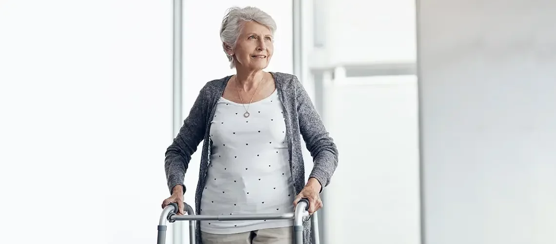 A smiling senior woman in a gray sweater walking with a short walker. For seniors, walkers are often critical.