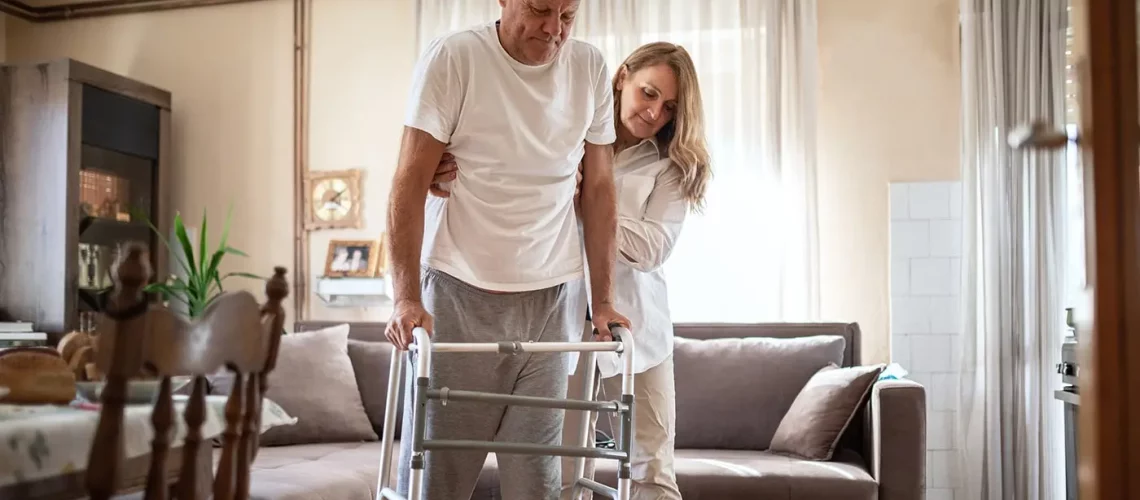 A tall senior trying to walk in his living room with the help of a walker while a caregiver is assisting him.