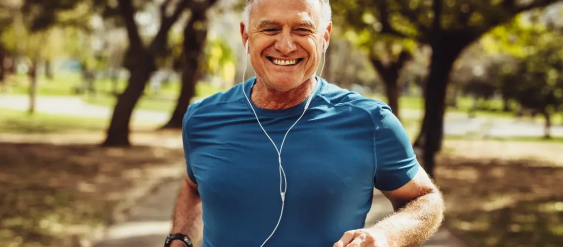 A smiling older adult man running in the park with earphones on.
